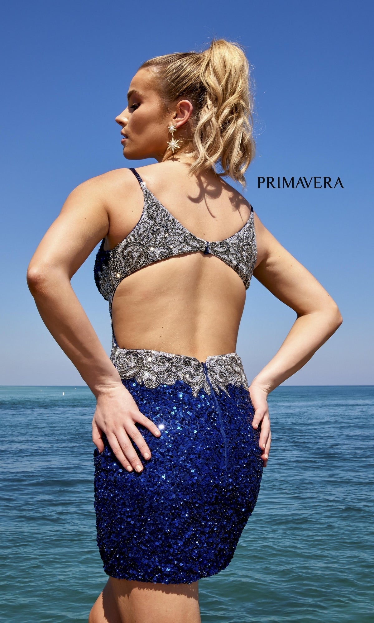 A blonde woman with a ponytail stands by the ocean wearing the Side-Beaded Short Sequin Party Dress 4052 in blue and silver with an open back.
